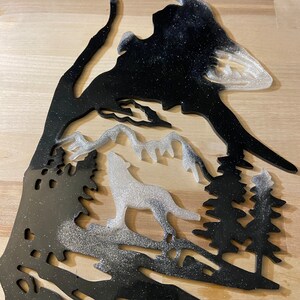 Puede incluir: Una decoración de pared de resina negra y blanca con purpurina que representa un lobo aullando a la luna con una cordillera y pinos en el fondo.