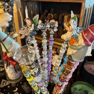 Può includere: Una collezione di bacchette perlate colorate con punte di cristallo. Le bacchette sono esposte in un vaso di vetro trasparente. Le bacchette sono decorate con varie perline, tra cui perline trasparenti, bianche, viola, blu e arancioni. Le bacchette sono sormontate da punte a forma di cristallo.
