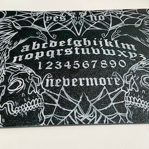 Puede incluir: Una tabla de Ouija en blanco y negro con un diseño de telaraña, dos cuervos y dos calaveras. La tabla presenta las letras del alfabeto, los números del 1 al 9 y el 0, y las palabras "sí", "no" y "nunca más".