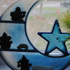 May include: A blue and white crescent moon shelf with a blue star cutout. The shelf has three tiers and features three black silhouette figurines.