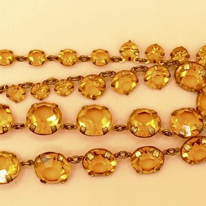 Pode incluir: Um colar de estilo vintage com várias fileiras de pedras preciosas redondas de cor âmbar. As pedras são engastadas numa corrente de metal dourado, com um fecho de gancho. O colar é exibido sobre um fundo bege claro.