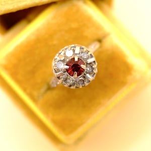 Può includere: Un anello con una pietra centrale rotonda rosso scuro circondata da un alone di pietre più piccole e chiare incastonate in una fascia metallica. Il design circolare a grappolo mette in risalto il contrasto tra il centro rosso e le pietre.