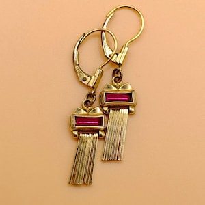 Może przedstawiać: Para złotych kolczyków z prostokątnym, rubinowym kamieniem i frędzlami z pionowych prętów. Kolczyki mają zapięcie zatrzaskowe i wygląd vintage. Prezentowane na brzoskwiniowym tle.