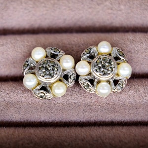 Puede incluir: Un par de pendientes de plata con un diseño floral. Cada pendiente presenta un círculo central de piedras de marcasita negras rodeadas de cinco perlas blancas.