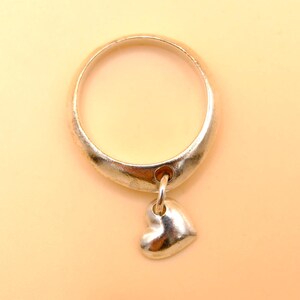 Op de afbeelding: Een zilverkleurige metalen ring met een gladde, gepolijste band in een licht taps toelopend ontwerp. Een kleine, bolvormige hartvormige bedel is aan de onderkant van de ringband bevestigd met een ronde ring, wat een bungelend element creëert.