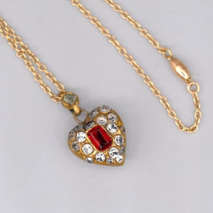 May include: A gold chain necklace with a heart-shaped pendant. The pendant is set with small clear stones and a larger red stone in the centre.
