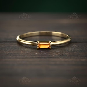Op de afbeelding: Een delicate gouden ring met een rechthoekige, oranje-gele edelsteen. De ring heeft een dunne, gepolijste band en de edelsteen is in een minimalistische stijl gezet. De ring wordt getoond op een donker houten oppervlak.