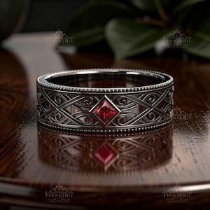 May include: A silver-toned metal band ring featuring an intricate openwork filigree pattern of swirling lines and geometric shapes. A single square-cut red gemstone is set centrally within the band, bordered by beaded edges along the top and bottom.