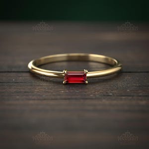 May include: A gold ring with a rectangular, deep red gemstone. The ring band is thin and polished, and the gemstone is set with four prongs. The ring is displayed on a dark wooden surface.