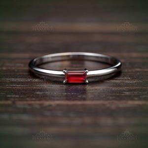 May include: A silver ring featuring a rectangular, vibrant red gemstone. The ring has a slender band and a minimalist design, showcasing the gemstone's color and shape. The ring is set against a dark wood grain background.