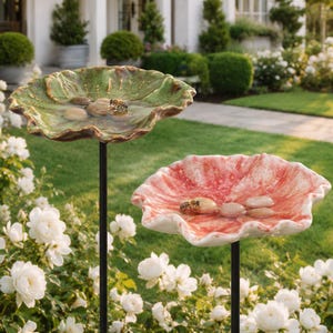 May include: Two ceramic bird baths on thin black metal stands, one with a green glazed finish and the other with a red and white mottled finish. Each shallow basin contains water and smooth river stones, with a honeybee resting on the stones.