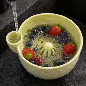 May include: A pale yellow ceramic berry colander with a side handle and a central drainage dome. The bowl is filled with water, fresh red strawberries, and dark blue blueberries, with a stream of water flowing into the side handle.