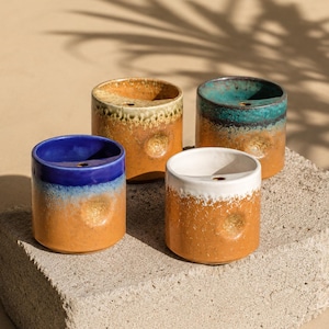 May include: Four small, cylindrical ceramic planters with a textured finish. Each planter has a different color scheme: blue and orange, tan and orange, teal and orange, and white and orange. The planters are displayed on a concrete surface.