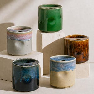 Op de afbeelding: Vijf cilindervormige keramische vaten met een halfronde opening aan de bovenkant en een klein rond gat. De vaten zijn voorzien van diverse reactieve glazuren in effen groen, amberbruin en tweekleurige combinaties van blauw en crème.