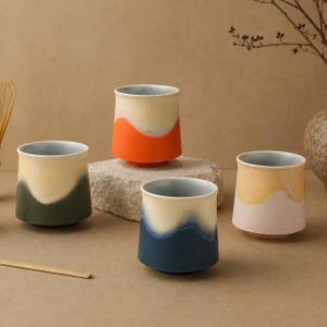 Op de afbeelding: Vier keramische theekopjes met een uniek ontwerp. Elk kopje heeft een ander kleurenschema, waaronder oranje, groen, blauw en roze, met een lichtgekleurd interieur. De kopjes hebben een moderne, minimalistische uitstraling.