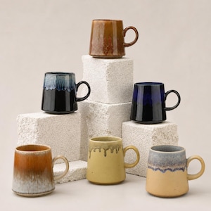 May include: A collection of six ceramic mugs in various colors, including brown, blue, yellow, and gray. Each mug has a unique glaze and a rounded handle. The mugs are arranged on white, textured blocks, creating a visually appealing display.