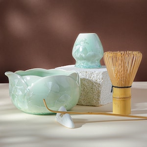 Può includere: Una ciotola per matcha in ceramica verde chiaro con bordo smerlato e motivi a foglia di ginkgo in rilievo, un supporto per frusta verde abbinato, una frusta da tè in bambù con filo nero e un lungo cucchiaio da tè in bambù su supporto.