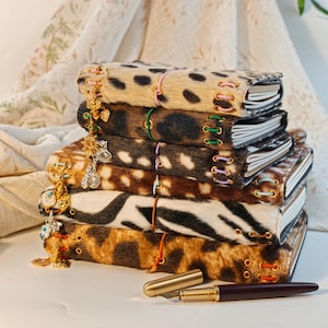 May include: A stack of five journals with textured animal print covers, including leopard, cheetah, and zebra patterns. Each journal is bound with colorful elastic cords and metal eyelets, featuring dangling gold-tone charms.