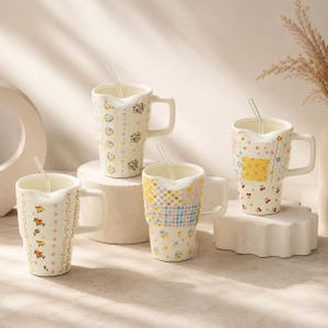 May include: Four white ceramic mugs with floral and patchwork designs, each with a clear spoon. The mugs have a unique wavy rim and square handles. The mugs are arranged on a white surface.