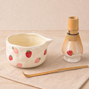 Puede incluir: Un cuenco de matcha de cerámica color crema, decorado con fresas rojas y rosas. También hay un batidor y una cuchara de bambú. El batidor tiene un diseño de fresa en la base. Los artículos están sobre una superficie de lino beige.