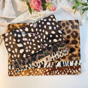 May include: A stack of rectangular, animal-print clutches with fur-like texture. Each clutch features gold-toned personalized names, small metallic charms, and decorative colored stitching details. The patterns include spotted and leopard prints.