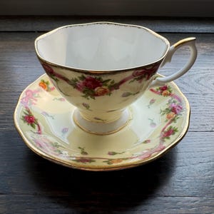 May include: A yellow teacup and saucer set with a floral pattern of pink, red, and yellow roses and pink ribbons. The set has a gold trim.