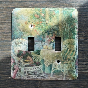 May include: A decorative light switch cover featuring a vintage garden scene. The cover depicts a patio with wicker chairs, a table, and floral arrangements. The color palette includes greens, pinks, and yellows, creating a charming, antique aesthetic.