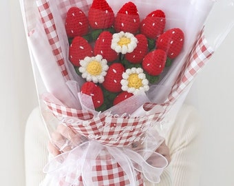 May include: A bouquet of crocheted strawberries with white flowers, wrapped in red and white checkered paper and tied with a white ribbon. The bouquet is held by a person wearing a white sweater.