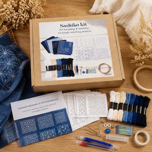 May include: A Sashiko mending and marking kit in a cardboard box featuring 14 simple stitching projects. The kit includes white, cream, and blue embroidery threads, white plastic pattern templates, needles, a small blue label, and two pens.