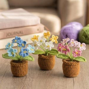 Op de afbeelding: Drie gehaakte bloemstukken in bruine potten. De bloemen zijn blauw, geel en roze, met groene bladeren. De potten staan op een houten oppervlak. Een boek met het woord "JOURNAL" staat op de achtergrond.