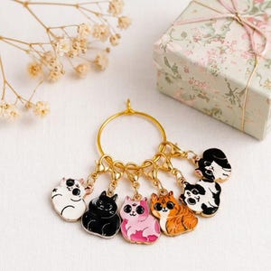 May include: A gold-toned metal ring featuring six dangling enamel cat charms. The charms include a white cat, a solid black cat, a pink cat, an orange tabby cat, a black and white cat, and a black and white cat with a different pattern.