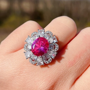 Może przedstawiać: Srebrny pierścionek z dużym, okrągłym, różowym kamieniem szlachetnym otoczonym mniejszymi, przezroczystymi kamieniami szlachetnymi. Pierścionek jest na ręce z zegarkiem na nadgarstku.