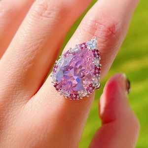 Może przedstawiać: Pierścionek z różowym kamieniem szlachetnym w kształcie gruszki, otoczony halo mniejszych różowych i białych kamieni szlachetnych. Pierścionek jest osadzony w srebrze i noszony na ręce.