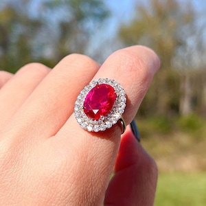 May include: A silver ring with a large oval-shaped red gemstone surrounded by a double halo of smaller clear gemstones.