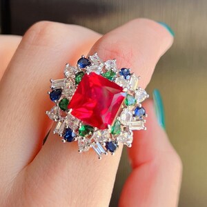 Könnte beinhalten: Ein silberner Ring mit einem großen quadratischen roten Edelstein, umgeben von kleineren weißen, grünen und blauen Edelsteinen.