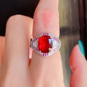 Może przedstawiać: Srebrny pierścionek z dużym, kwadratowym, czerwonym kamieniem szlachetnym jako centralnym elementem. Pierścionek jest ozdobiony mniejszymi, prostokątnymi i okrągłymi kamieniami w kolorze białym i różowym, tworzącymi uderzający kontrast. Pierścionek jest noszony na palcu.