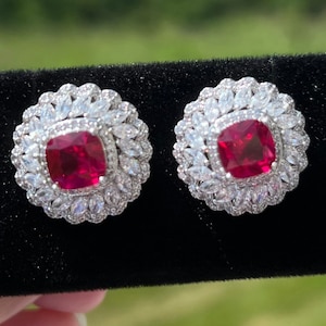 May include: A pair of silver earrings with a red gemstone in the centre of each earring. The earrings are surrounded by smaller clear gemstones.