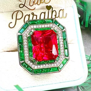 May include: A red gemstone ring with a square emerald cut. The ring is set in a silver band with a halo of smaller green gemstones and diamonds.
