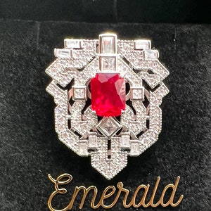 以下が含まれることがあります： 赤い正方形の大きな宝石が、小さな透明な宝石で囲まれた銀の指輪。指輪は、金色の文字で「Emerald Love Paraiba」と書かれた黒いベルベットの箱に入っています。