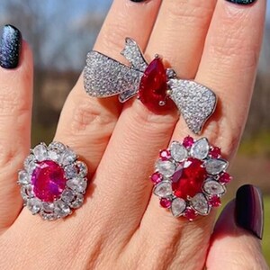 Könnte beinhalten: Drei silberne Ringe mit roten Edelsteinen und Diamantbesatz. Die Ringe befinden sich an einer Hand mit dunkelgrünem Nagellack.