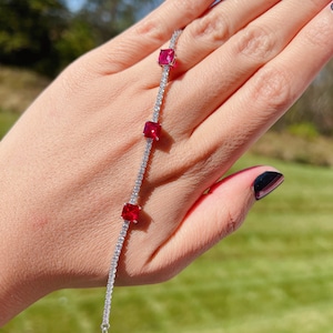 Pode incluir: Um bracelete de prata com uma corrente delicada e três pedras preciosas quadradas vermelhas. O bracelete está no pulso de uma pessoa.