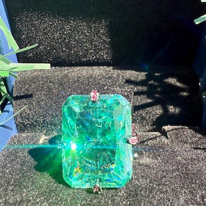 May include: A large, emerald-cut gemstone ring with a vibrant green hue. The ring features a rectangular stone with multiple facets, accented by smaller pink gemstones. The word "Paraiba" is displayed in gold script above the ring.