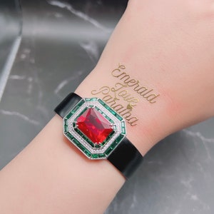 Puede incluir: Un brazalete de cuero negro con una gran piedra roja rodeada de piedras verdes más pequeñas. El brazalete tiene un cierre plateado y el texto "Emerald Love Paradise" está escrito en oro en la muñeca.