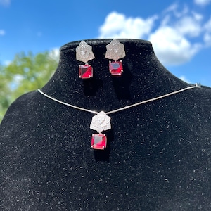 May include: A silver necklace and earrings set with a red gemstone pendant and matching earrings. The pendant and earrings feature a floral design with clear cubic zirconia stones.