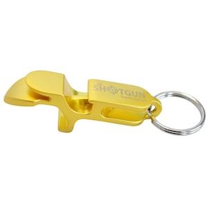May include: A gold-colored bottle opener with a key ring. The opener has the text "THE SHOTGUN" and a target symbol. The design is for opening bottles. The opener is made of metal.