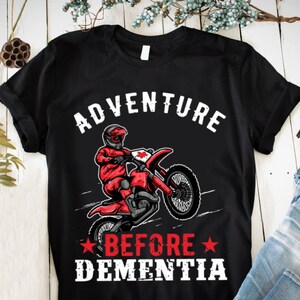 May include: Black t-shirt with a white graphic of a person riding a dirt bike with the text "Adventure Before Dementia" in white.