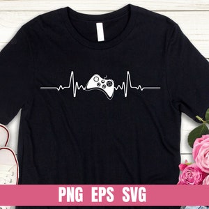 Puede incluir: Camiseta negra con un gráfico blanco de un mando de videojuego con una línea de latido del corazón. El texto "PNG EPS SVG" está en la parte inferior de la camiseta.