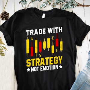 Op de afbeelding: Zwart T-shirt met een witte afbeelding met de tekst "Trade with Strategy, Not Emotion". De afbeelding bevat een candlestick-grafiek met valuta symbolen zoals dollar, pond, yen en euro.
