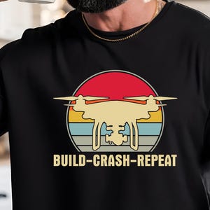 May include: Black t-shirt featuring a retro-style drone graphic inside a circular design with red, orange, yellow, and blue stripes. The text "BUILD-CRASH-REPEAT" is below the drone image.