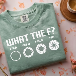 Puede incluir: Una camiseta verde salvia con la frase "WHAT THE F?" en letras blancas. Debajo, símbolos de apertura y números f/1.8, f/2.8, f/5.6 y f/8. La camiseta doblada está sobre una superficie con estampado floral. Marca Comfort Colors.